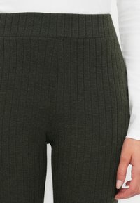 Olijfgroene geribbelde gebreide leggings hebben een hoge tailleband en een zachte textuur. De stof toont verticale strepen en heeft een aansluitende pasvorm.