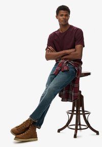 Donkerrode T-shirt met ronde hals, blauwe spijkerbroek, rood-zwarte geruite blouse geknoopt rond de taille en bruine veterschoenen met gele veters.