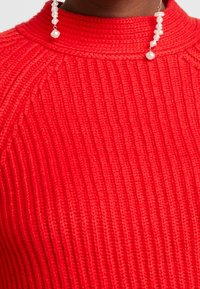 Červený žebrovaný svetr s vysokým límcem. Má pletenou texturu s výraznými svislými liniemi. Jsou viditelné náušnice s nepravidelnými perlovými detaily.