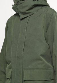 Veste imperméable verte en tissu texturé à rayures, avec un col montant, des poches avant et une capuche ajustable.