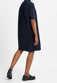 Marineblaues Hemdblusenkleid mit kurzen Ärmeln, einem geknöpften Kragen und einer lockeren Passform, getragen mit schwarzen Loafers. Glatte Stoffstruktur.