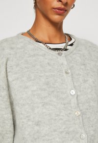 Light gray cardigan made from soft material, featuring a button-up front with four round buttons, and a crew neckline over a striped shirt.