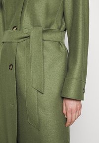 Oliivivihreä takki, joka on valmistettu teksturoidusta kankaasta ja jossa on solmittava vyö, suuret napit ja muotoillut hihansuut. Yksityiskohtainen ompelu saumoissa.
