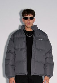 Kurtka puchowa w ciemnoszarym kolorze, z kołnierzem, zapięciem na napy z przodu oraz kieszeniami po bokach. Nosi ją osoba w czarnej koszuli; model uzupełnił look okularami przeciwsłonecznymi.