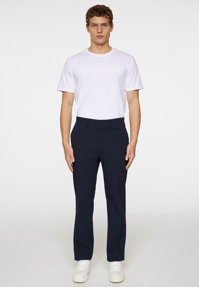 Meripaka dressia navy dengan potongan yang disesuaikan, depan datar, dan penutupan dengan tombol tunggal, dipasangkan dengan kaos putih leher bulat polos dan sepatu sneakers putih.