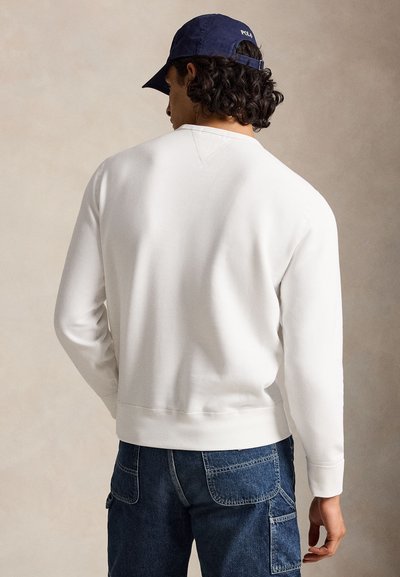 Mand iført en hvid langærmet sweatshirt, mørkeblå denimjeans med lommer og en navyblå baseballkasket vendt bagud.