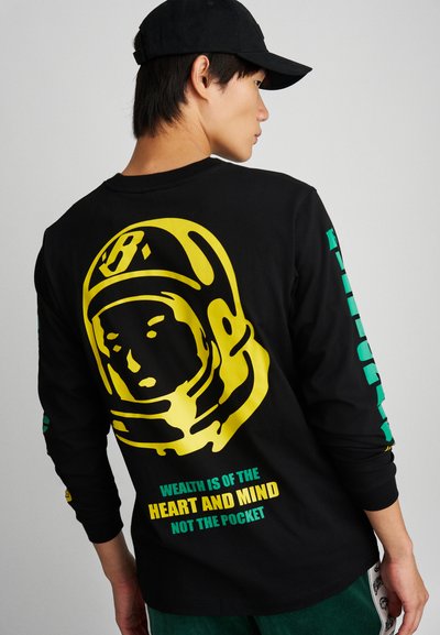 Camiseta negra de manga larga con un gran gráfico de un astronauta amarillo en la parte posterior. El texto debajo del gráfico dice: "LA RIQUEZA ES DEL CORAZÓN Y LA MENTE, NO DEL BOLSILLO."
