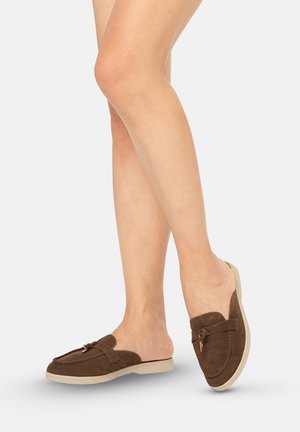 Bruine suède instappers met een decoratieve kwast, gedragen op blote voeten tegen een effen lichte achtergrond.