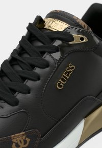 Zapatillas negras con acabado de cuero suave, detalles en dorado, cordones tono sobre tono y panel lateral estampado que presenta un logotipo en marrón y dorado.
