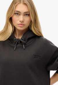 Sweat-shirt à capuche noire avec cordon de serrage, arborant un logo brodé sur la poitrine. Le tissu semble doux et texturé.