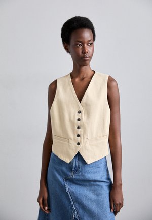 Beige linen waistcoat with a v-neck, five black buttons, and two front pockets, worn over a denim skirt with a frayed hem.