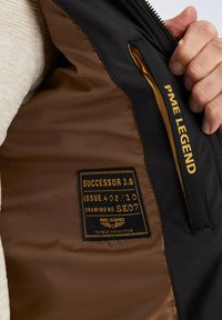 Men's bomber jacket interior with brown lining, black outer shell, stitched tag labeled "SUCCESSOR 3.0," and yellow logo accents.