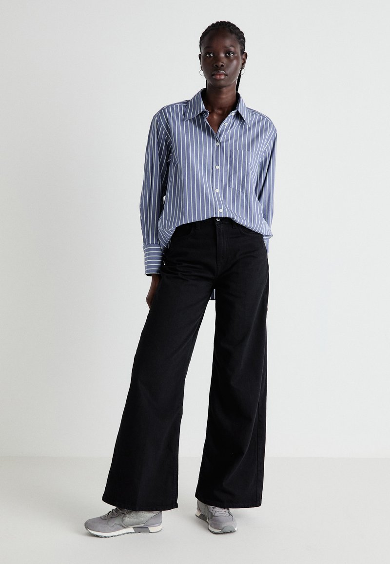 Camicia a righe blu e bianche con bottoni, pantaloni larghi neri a gamba larga e scarpe sportive grigie. Il modello è in piedi con le mani in tasca contro uno sfondo neutro.