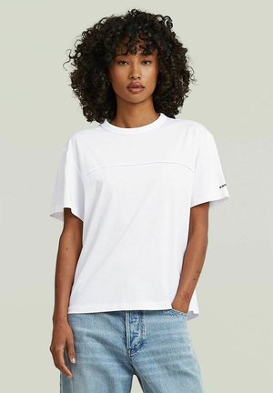 Donna con capelli ricci che indossa una t-shirt bianca semplice a maniche corte e jeans azzurri, in piedi con una mano in tasca.