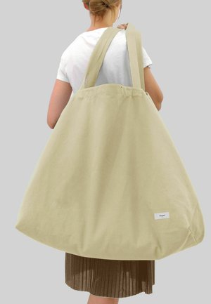Stor, beige kanvas toteväska med långa handtag, mjuk textur och rundad botten. Har en liten vit logolapp på ena sidan.