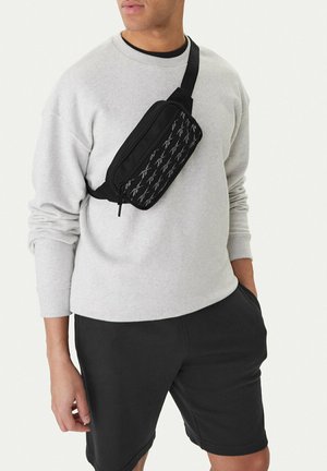 Persona che indossa una felpa grigio chiaro e pantaloncini neri con una borsa a tracolla nera con una tasca frontale decorata.