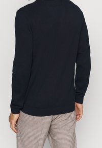 En person som bär en marinblå tröja med lång ärm, vänd bort, kombinerad med rutiga byxor. Bakgrunden är enfärgad och ljus.