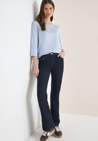 Femme appuyée contre un mur blanc portant un haut à manches longues bleu clair, un jean bleu foncé et des baskets marron avec des lacets blancs.