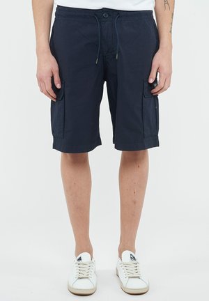 Uomo che indossa pantaloncini cargo blu navy con coulisse, scarpe da ginnastica bianche e una maglietta bianca, in piedi davanti a uno sfondo uniforme.