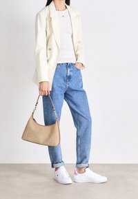 Beżowa torebka o gładkiej fakturze i srebrnych elementach. Biała dzianinowa bluzka, kremowa marynarka i jasnoniebieskie spodnie z wysokim stanem połączone z białymi trampkami.