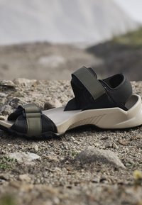 Pojedyncza czarna i oliwkowo-zielona sandałka outdoor z grubą amortyzowaną podeszwą spoczywająca na kamienistym podłożu.