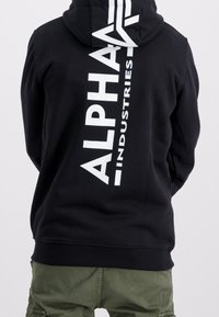 Felpa nera con cappuccio, con una stampa verticale bianca che riporta la scritta "ALPHA INDUSTRIES" sul retro. Il materiale appare liscio e morbido.