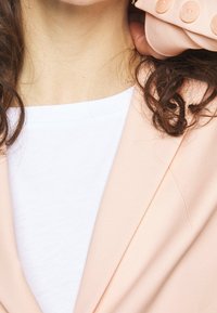 Marynarka z miękkiego, brzoskwiniowego materiału o gładkiej fakturze, zdobiona dekoracyjnymi guzikami i o dopasowanym kroju. Pod spodem znajduje się biały top z krótkim rękawem.