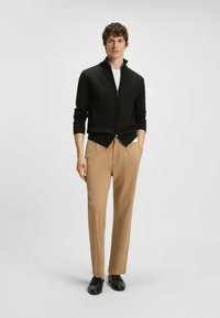 Svart zip-up kofta med ribbade ärmmuddar, matchad med beige byxor och svarta skor, stående mot en neutral bakgrund.