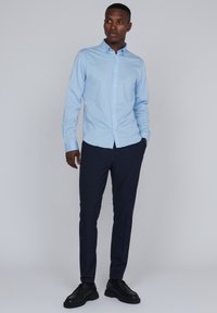 Chemise à manches longues bleu clair avec un col boutonné, associée à un pantalon bleu marine et des chaussures noires, sur un fond neutre.