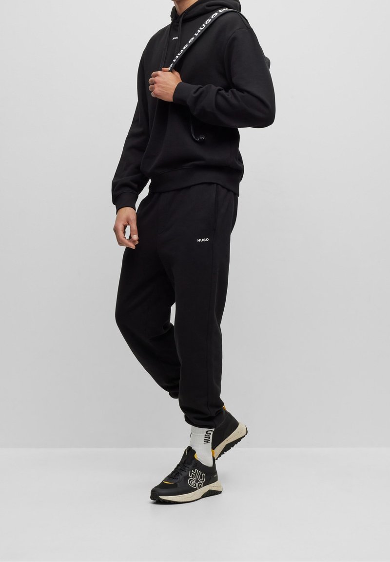 Personne portant un sweat à capuche noir Hugo et un pantalon de jogging, tenant une sangle d'épaule de marque, avec des baskets noires et jaunes et des chaussettes blanches avec logo.