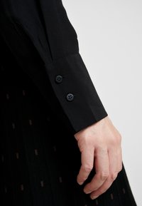 Manche de chemise noire à boutons aux poignets. Texture lisse, coupe ajustée. Main reposant sur une jupe plissée noire avec des motifs subtils.