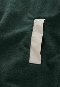 Fleece-kangas tumman vihreänä, jossa on kontrastivärinen valkoinen pehmeä paneeli. Korkeapintainen pinta, erottuvat saumat ja epäsäännöllinen muoto.