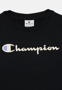 Črna bombažna majica s kratkimi rokavi in okroglim izrezom. Ima kremno belo "Champion" logotip s modrim akcentom.