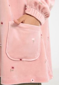 Indumento in pile rosa con motivi floreali. Presenta una tasca con bordi arrotondati e polsini elasticizzati, che mette in mostra una consistenza morbida.