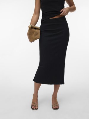 Woman wearing a black sleeveless top and black midi skirt, holding a tan clutch purse, and wearing braided strap open-toe sandals.