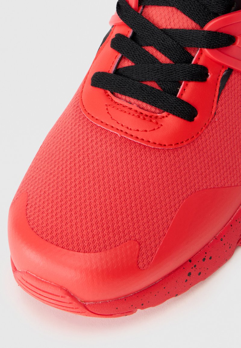 Chaussure de sport rouge avec une tige en mesh texturé, un embout en caoutchouc lisse et des lacets noirs. La semelle intermédiaire présente des accents à pois noirs.