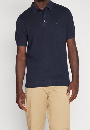 Uomo che indossa una polo blu navy a maniche corte e pantaloni beige, in piedi davanti a uno sfondo chiaro uniforme.