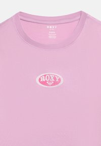 Λιλά βαμβακερό T-shirt με στρογγυλή λαιμόκοψη, τυπωμένο με λευκό και ροζ οβάλ λογότυπο που γράφει "ROXY" στο κέν τρο του στήθους.