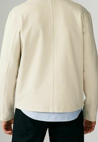 Veste beige texturée à coupe droite avec une patte de boutonnage dissimulée, portée sur une chemise rayée bleu clair et blanc.