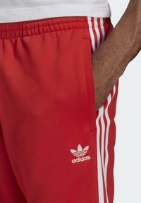 Pantaloni da jogging rossi Adidas con tre strisce bianche sui lati e logo Adidas, indossati con una maglietta bianca; mano in tasca.