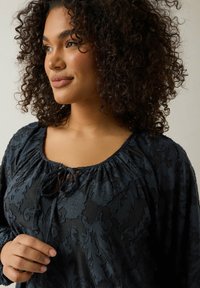 Schwarze Bluse mit floralen Mustern, versammeltem Ausschnitt und Bändern zum Binden. Der Stoff hat eine strukturierte Oberfläche und lange, luftig geschnittene Ärmel.