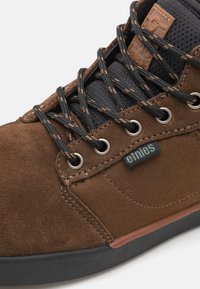 Brown suede shoe with black mesh upper, reinforced eyelets, contrasting brown stitching, and green logo tag. Rubber sole with slight accent.