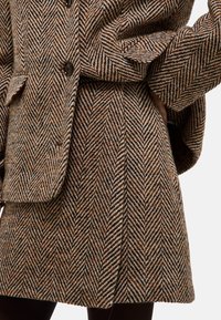 Manteau marron à motif chevron avec une coupe décontractée, de grands boutons et des poches à rabat. Tissu texturé, longueur genou avec des plis.