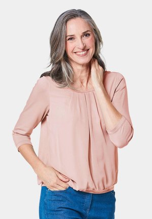 Pfirsichfarbene Bluse mit Dreiviertelärmeln, gefältelter Vorderseite, leichtem Stoff und einem lockeren, abgerundeten Saum. Kombiniert mit blauen Jeans.