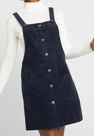 Kobieta ubrana w granatową sukienkę na szelkach z aksamitu sztruksowego z guzikami z przodu, na białym golfie z długim rękawem.