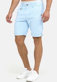 Pantalones cortos de algodón azul claro con dobladillos enrollados, cierre de botón y dos bolsillos laterales. Llevado con zapatillas blancas.