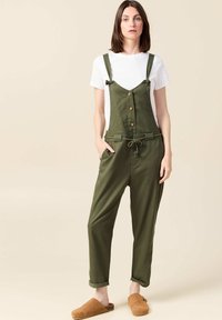 Olijfgroene overall met verstelbare banden, voorknopen en een trekkoord in de taille. Gecombineerd met een witte t-shirt en bruine slippers.