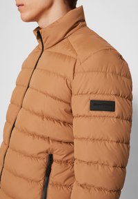 Giacca puffa marrone con colletto alto, cuciture orizzontali e una tasca con zip nera. Presenta un patch con il logo sulla manica sinistra.