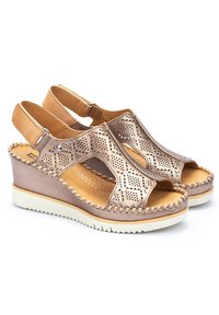 Sandalias de cuña con cuero metálico en rosa dorado perforado, punta abierta y correa trasera ajustable. Presenta una suela blanca de contraste y detalles trenzados.
