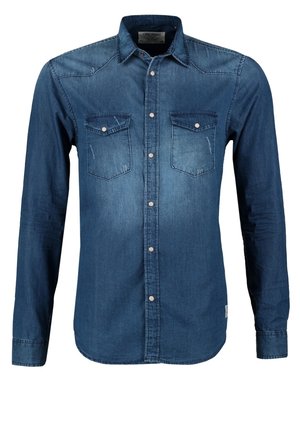 Mężczyzna w niebieskiej dżinsowej koszuli, czarnych spodniach, fioletowych trampkach i brązowych skórzanych szelkach idzie z jedną ręką w kieszeni.